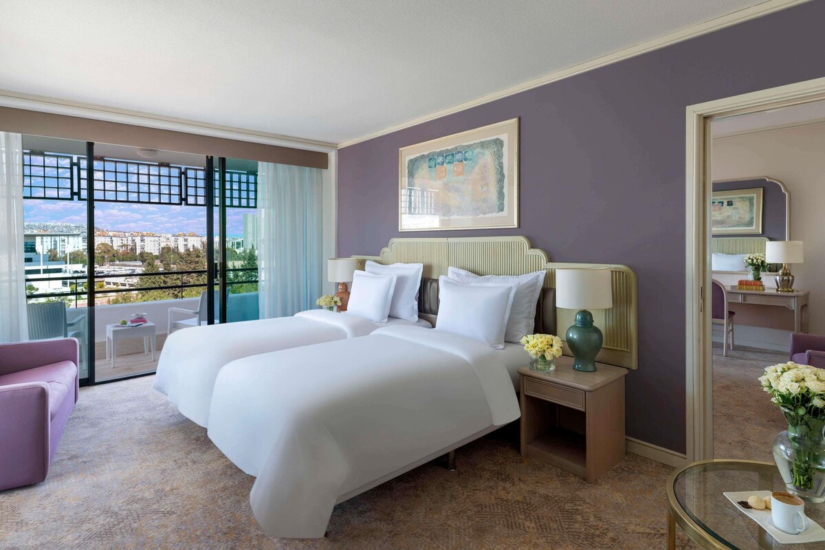 A hotel room with a large bed, two bedside tables, a chair, a glass table with a cup and cookies, and a balcony with a view of the city.