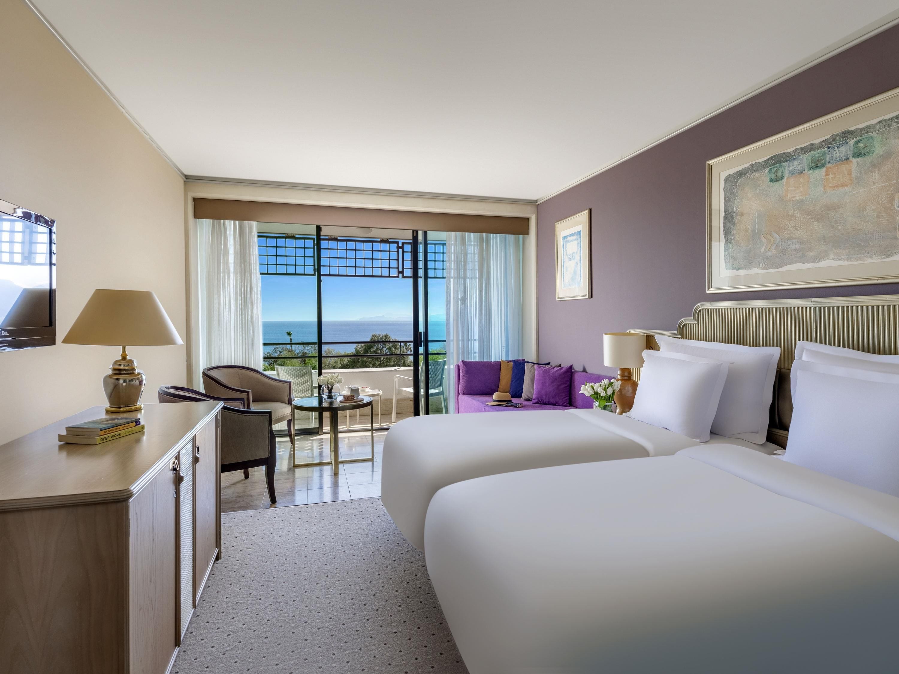Ein Hotelzimmer mit zwei Betten, einem Schreibtisch, einem Stuhl, einem kleinen Tisch und Blick aufs Meer.
