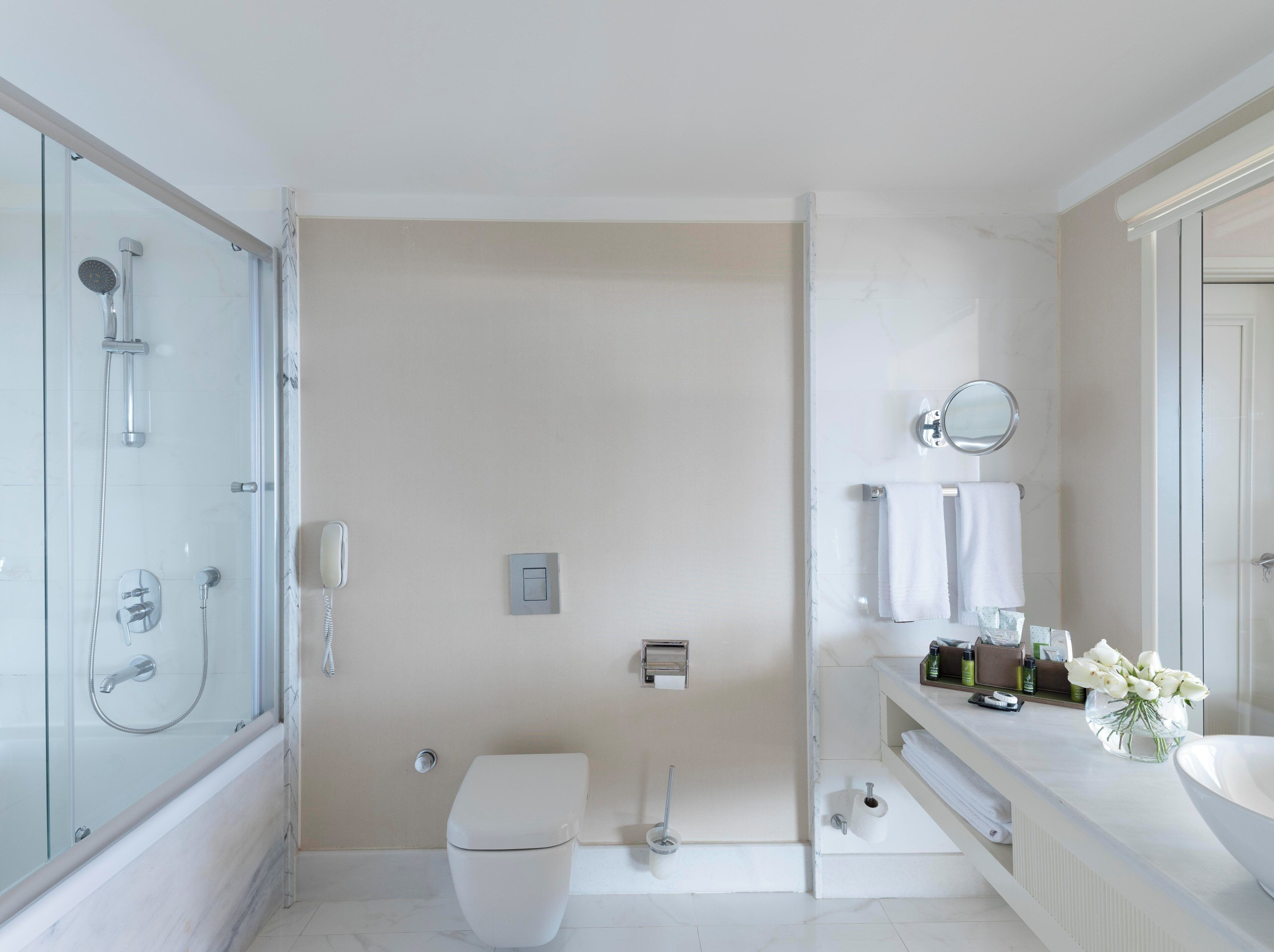 Un baño moderno con ducha de vidrio, inodoro, lavamanos y tocador con artículos de aseo.