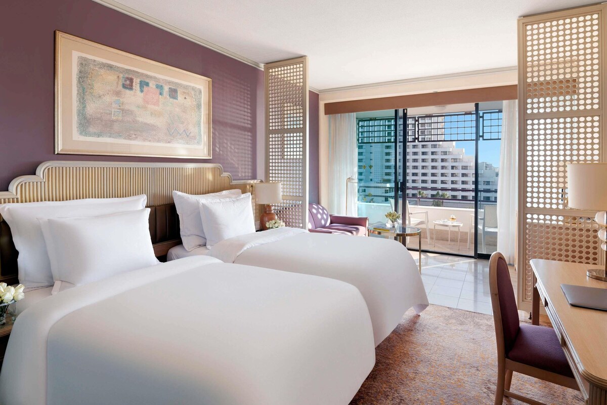 A hotel room with two beds, a large window with a city view, a dining area with a table and chairs, and a decorative wall panel.