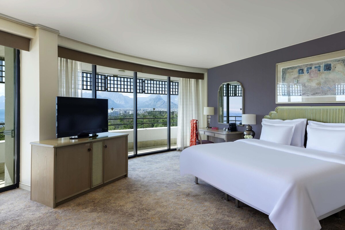 A hotel room with a large bed, a television on a wooden cabinet, a desk with a lamp, and a view of mountains through large windows.