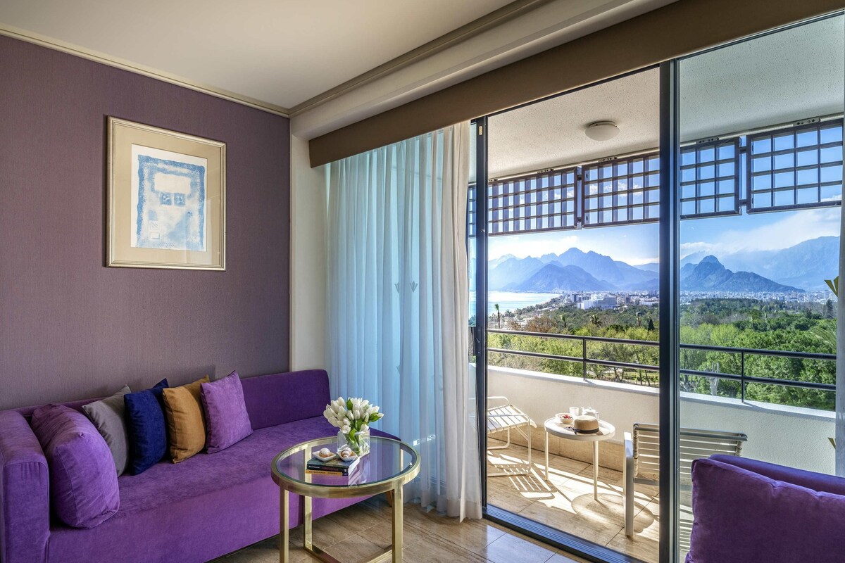A room with a purple sofa, a small round table with a vase of flowers, a framed picture on the wall, and a balcony with a view of mountains and a city.