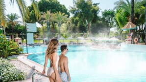 Una pareja de pie junto a una piscina, con palmeras y sillones de descanso al fondo.