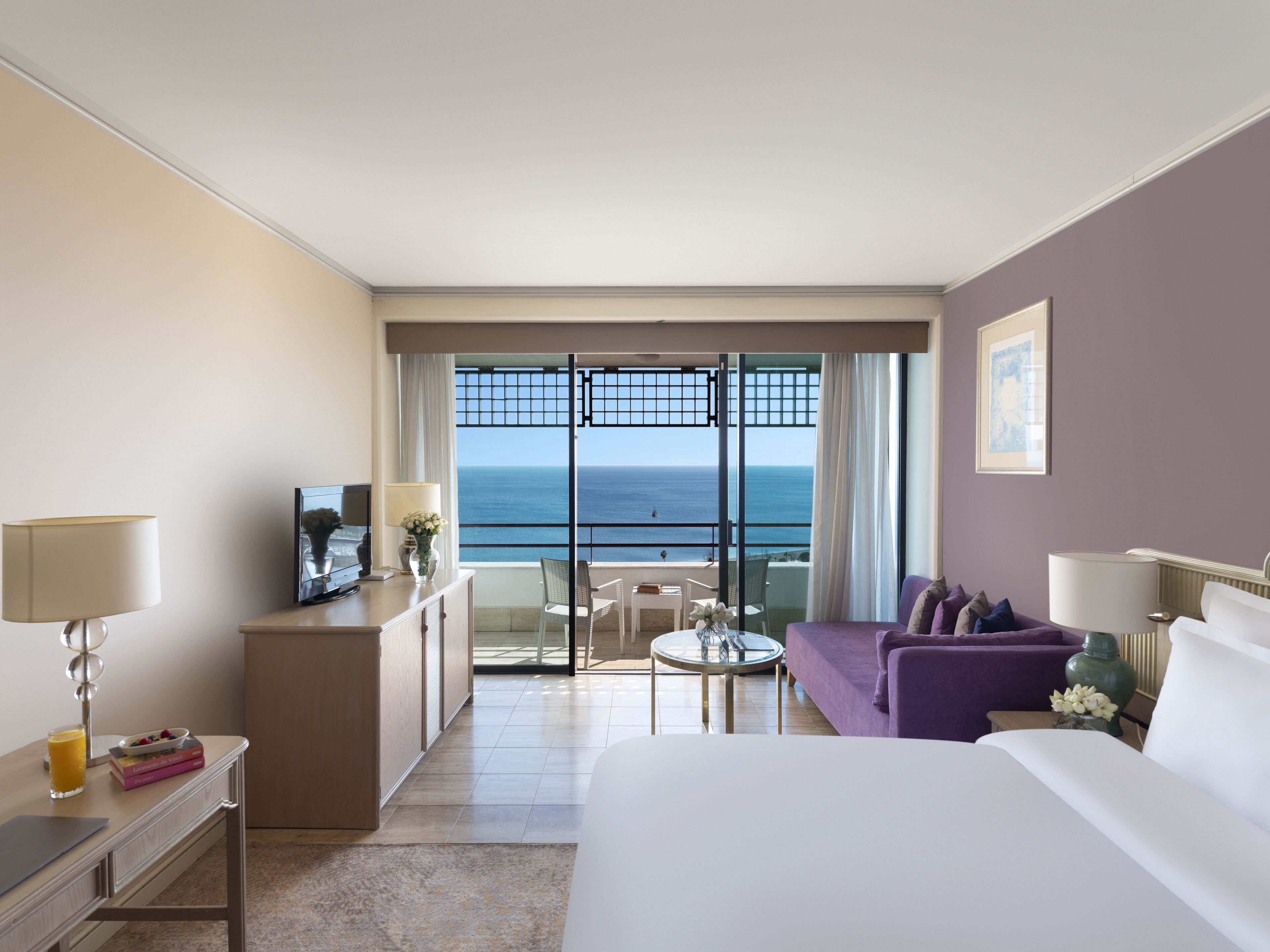 A hotel room with a bed, a desk, a chair, a sofa, a small table, and a view of the ocean through a balcony.