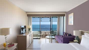 A hotel room with a bed, a desk, a chair, a sofa, a small table, and a view of the ocean through a balcony.