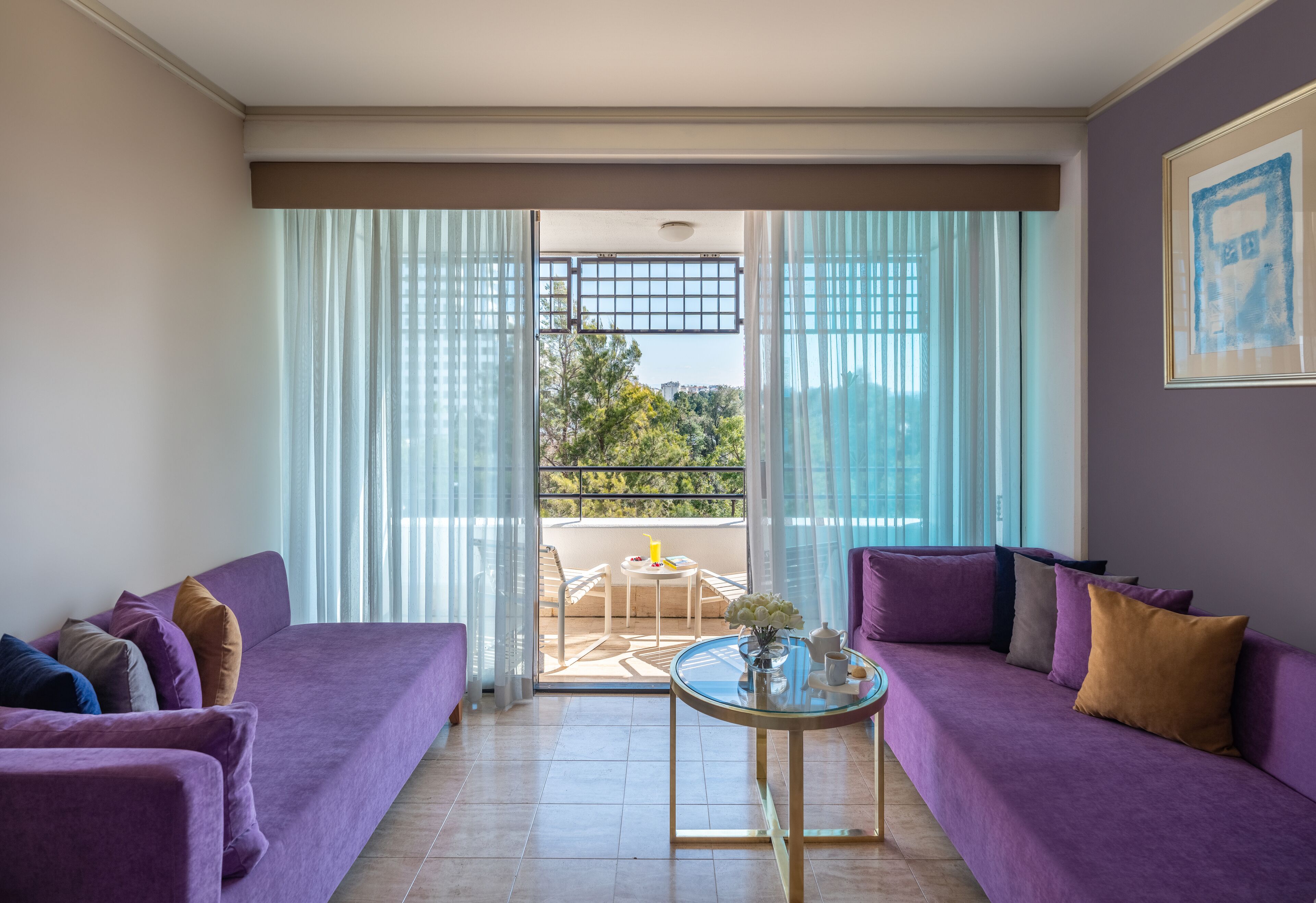 Un salón moderno con sofás morados, una mesa de centro de vidrio y un balcón con vistas a árboles y edificios.