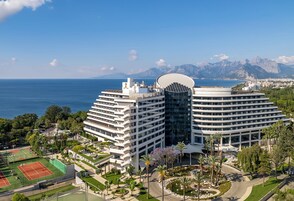 Un hotel moderno con cancha de tenis, rodeado de vegetación y vistas al mar y a las montañas.
