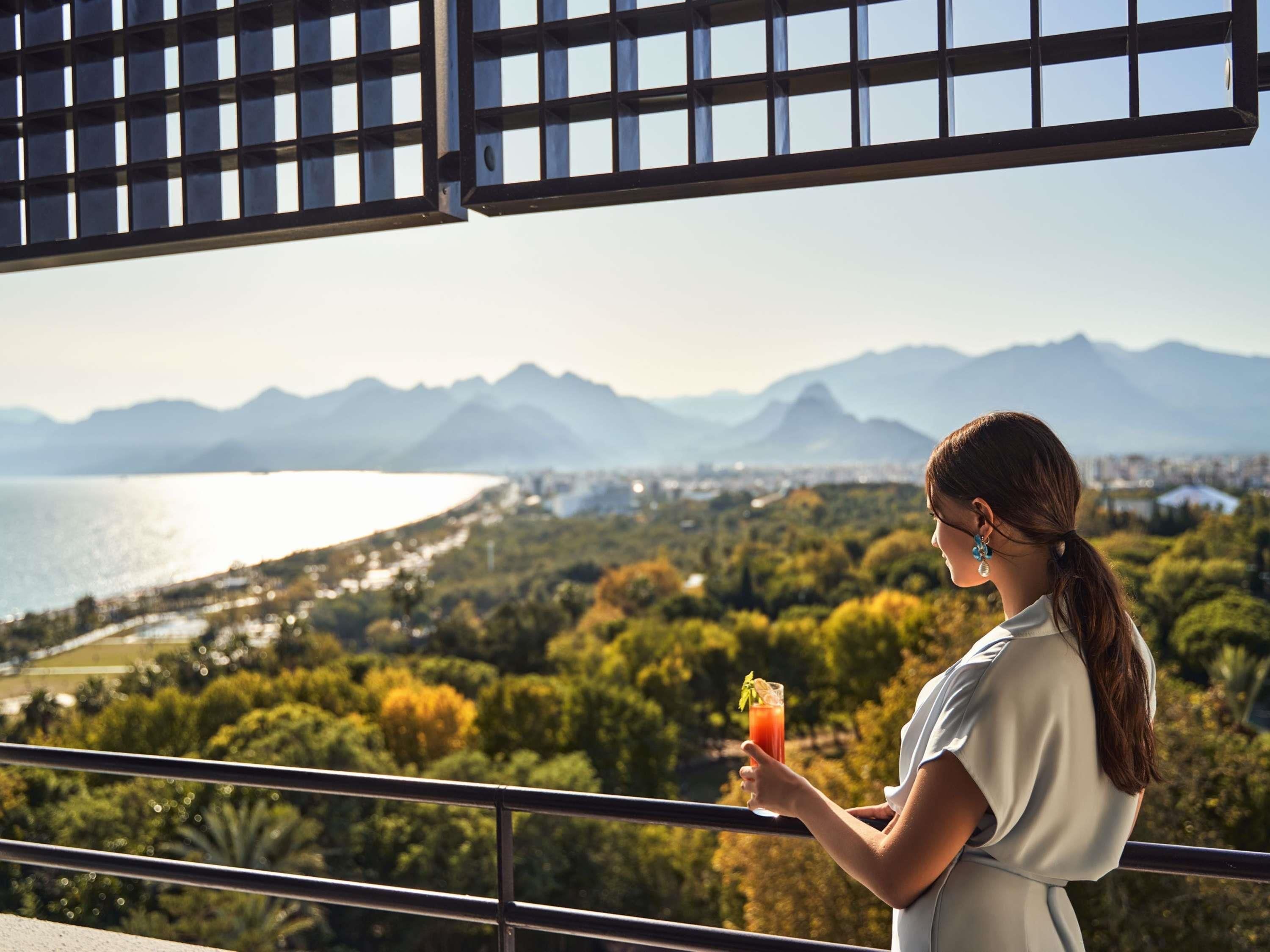 En person som håller i ett glas och blickar ut över ett vackert landskap med berg och en sjö eller ett annat vattendrag.