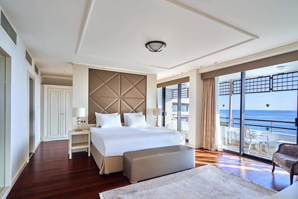 A modern hotel room with a large bed, a seating area, and a balcony with a view of the sea.