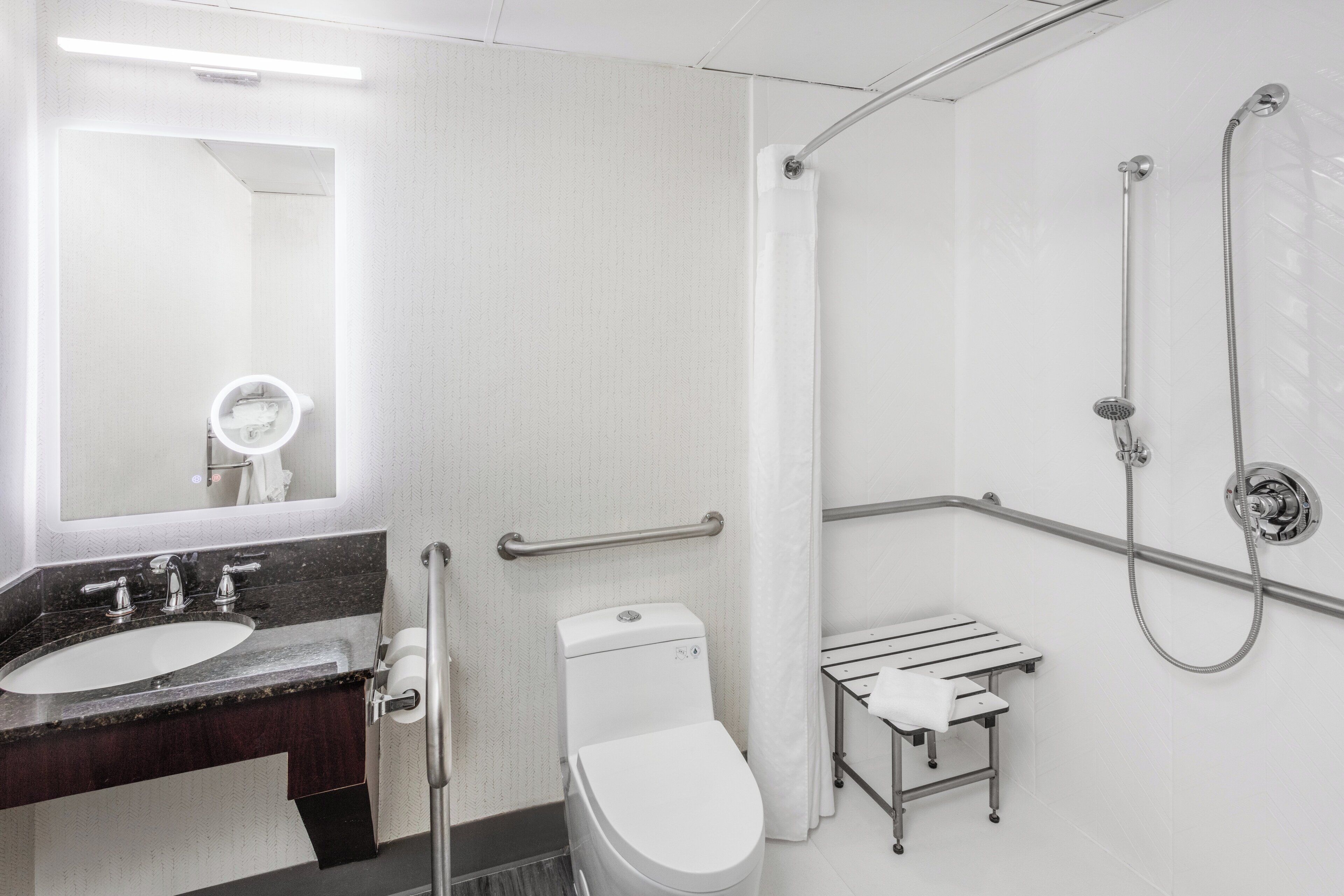 Une salle de bain moderne avec un lavabo blanc, un miroir et des toilettes, une douche équipée de barres d’appui et un petit porte-serviettes.