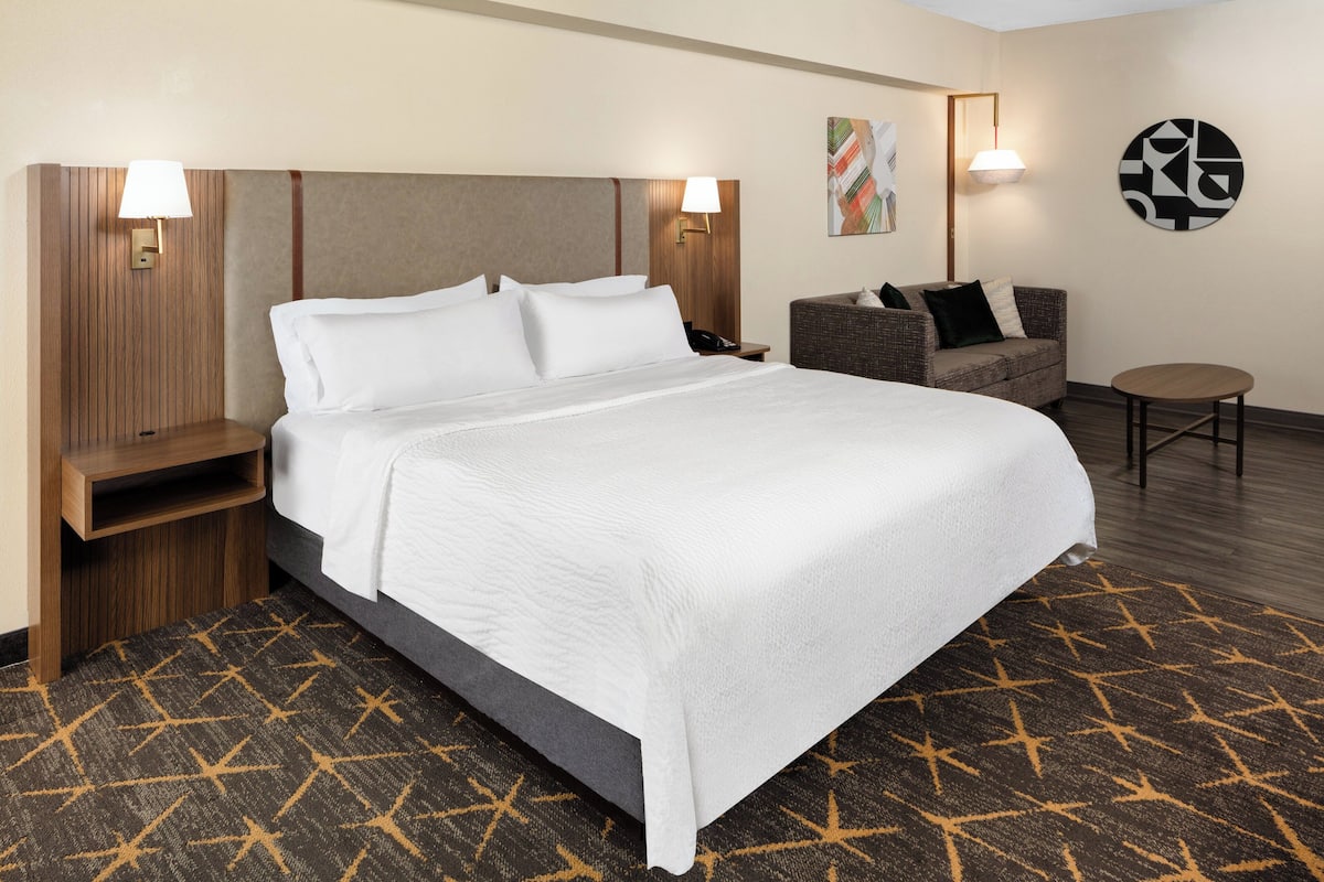 A hotel room with a large bed, a nightstand, a sofa, a small table, and two wall-mounted lamps.