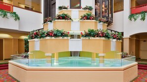 A hotel lobby with a central glass-bottomed pool, a staircase with red railings, and decorative plants.