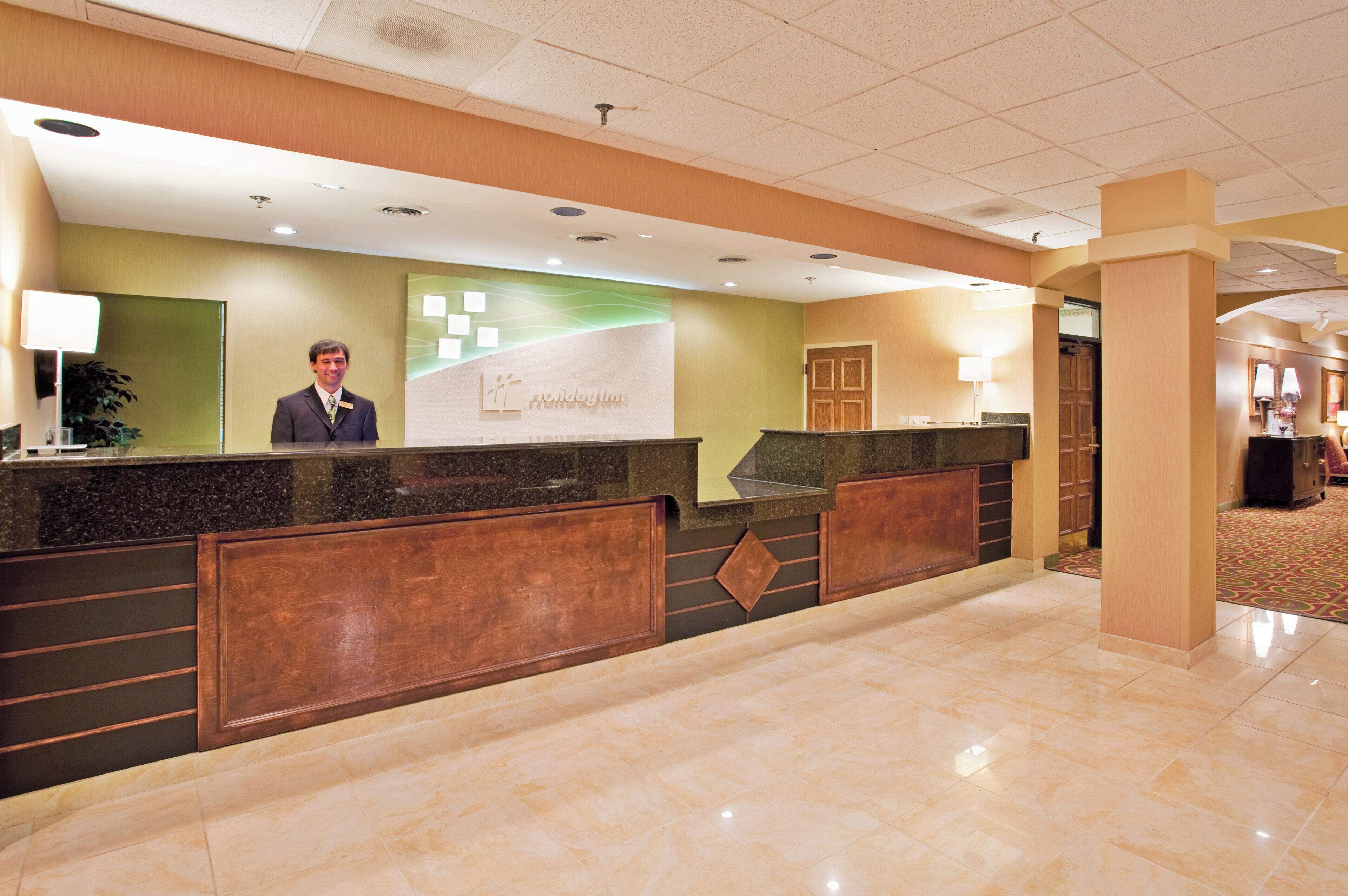 A hotel reception area with a receptionist, a reception desk, and a green wall with the hotel name.