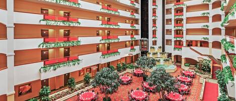 Un vestíbulo de hotel de varios niveles con suelos alfombrados de color rojo, varias mesas de comedor preparadas para los huéspedes, y balcones con barandillas rojas y vegetación.