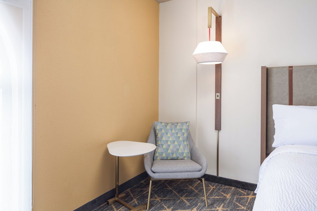 A hotel room with a bed, a small round table, a chair with a patterned cushion, and a floor lamp.