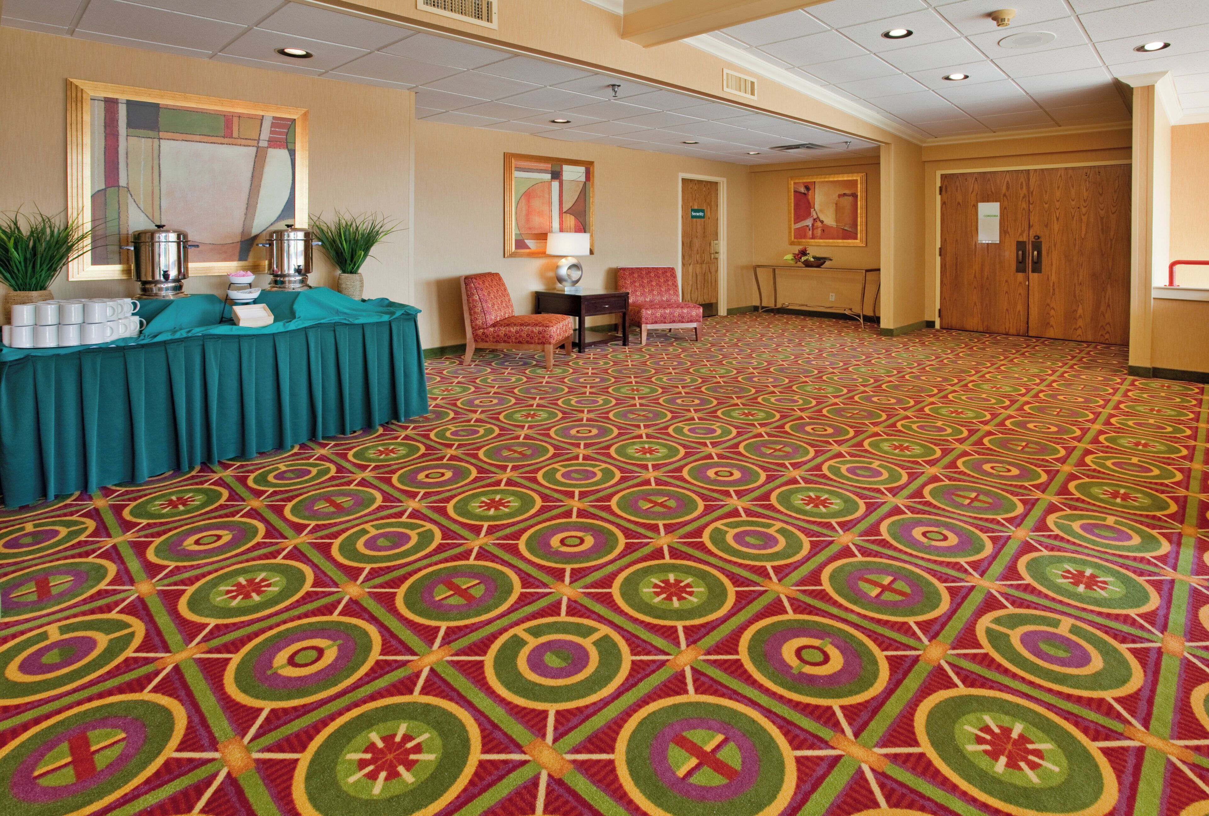 A hotel lobby with a patterned carpet, a reception desk, chairs, and a security door.