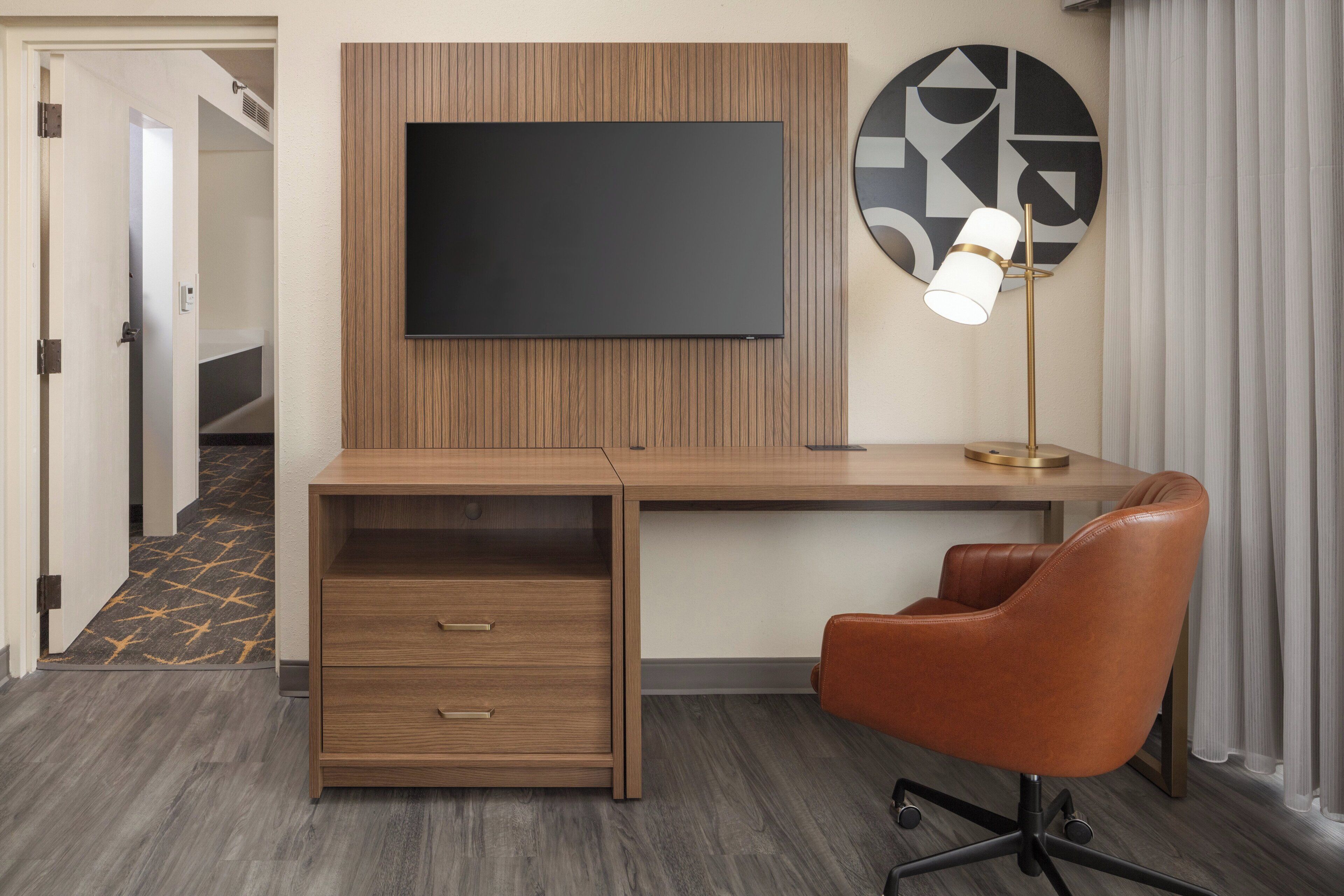 Une chambre d’hôtel avec un bureau en bois, un téléviseur à écran plat, une lampe moderne et un fauteuil en cuir marron.