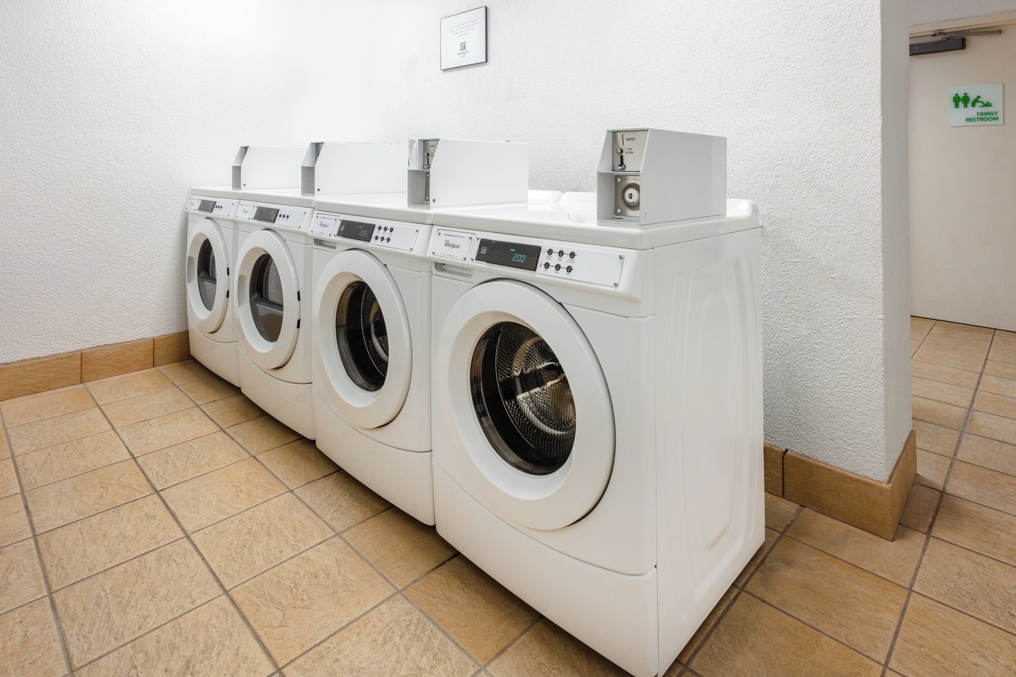 A row of white front-loading washing machines in a laundry room with a tiled floor and a sign indicating a family restroom.