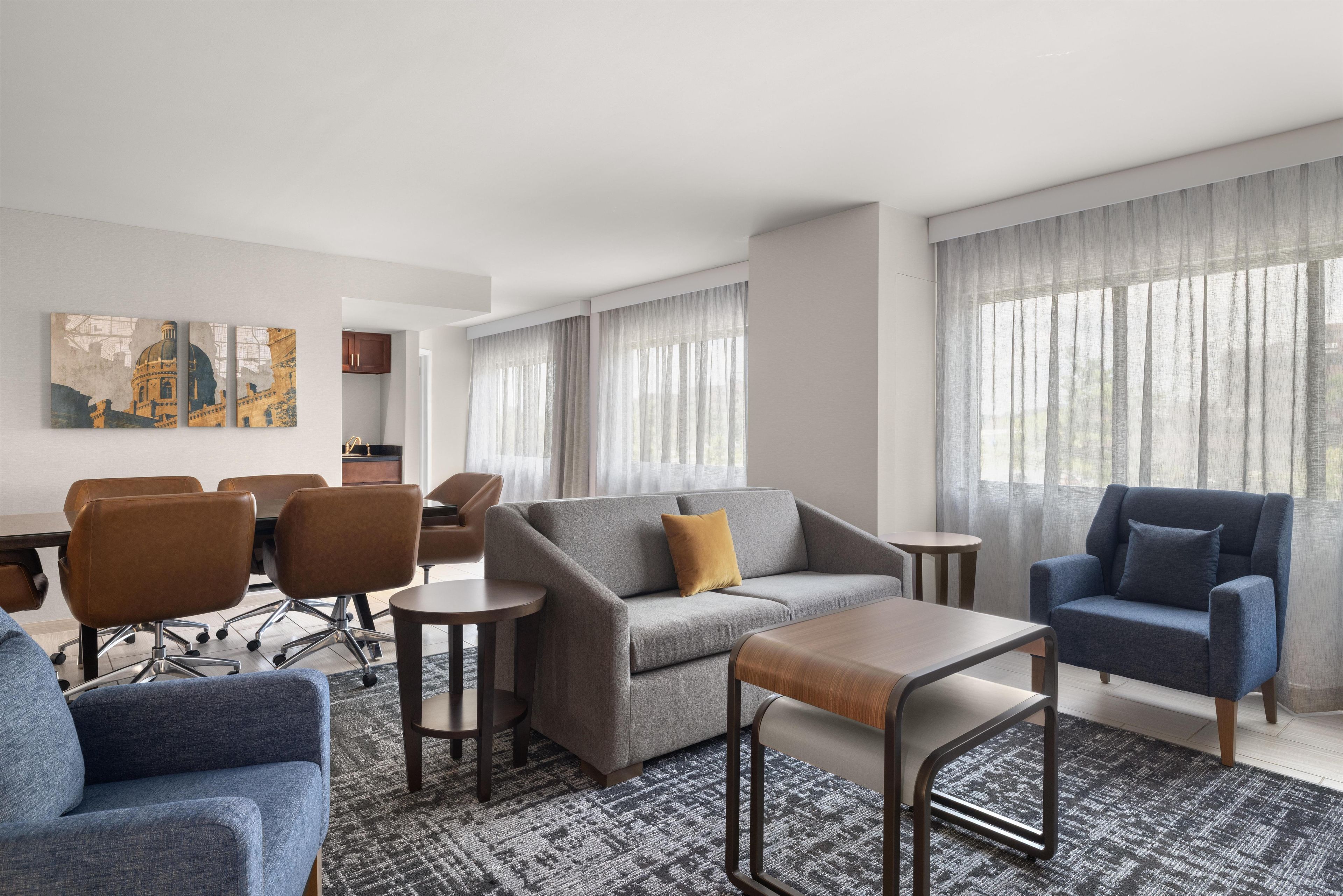 A modern hotel room with a grey sofa, blue armchairs, a small wooden table, and a dining area with chairs and a table.
