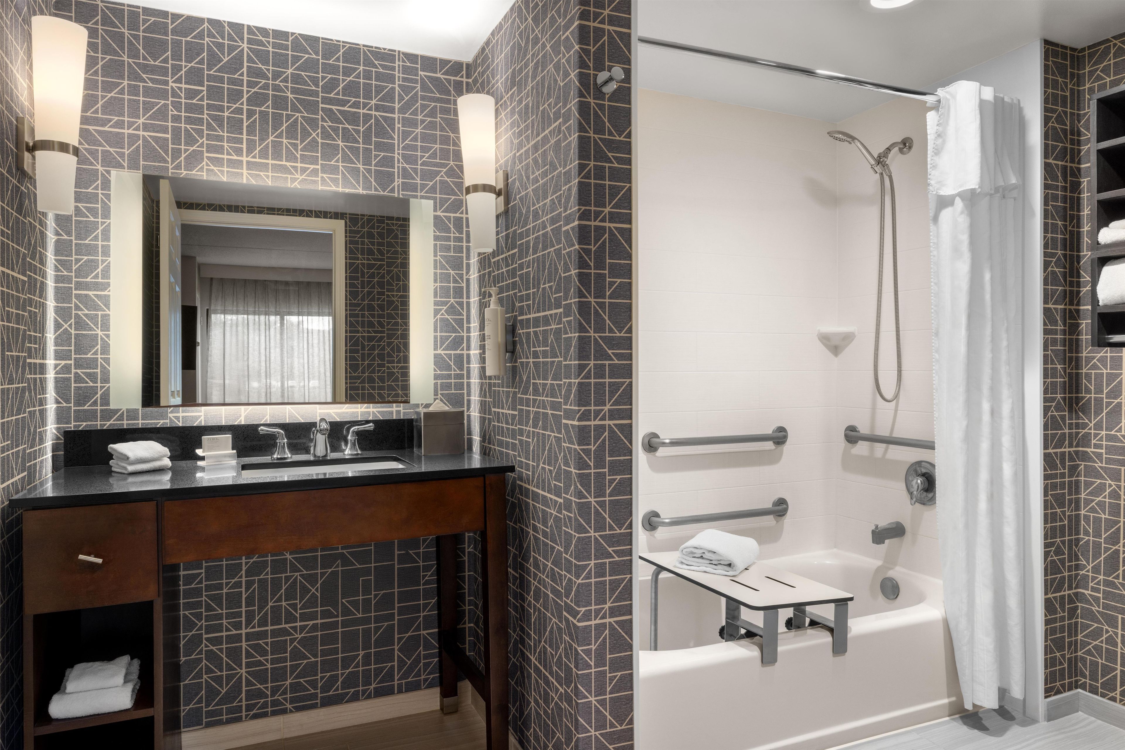 Un baño moderno con un espejo grande, un tocador con encimera oscura, una ducha con cabezal de ducha manual y una bañera.
