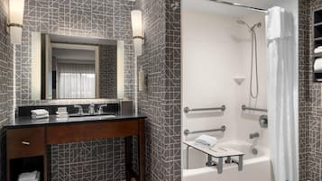 Un baño moderno con un espejo grande, un tocador con encimera oscura, una ducha con cabezal de ducha manual y una bañera.