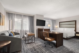 Habitación de hotel con cama, escritorio, silla, sofá, televisor y ventana con cortinas.