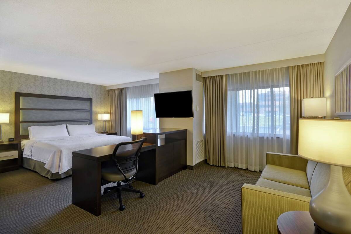 Een hotelkamer met een bed, bureau, stoel, televisie en grote ramen met gordijnen.