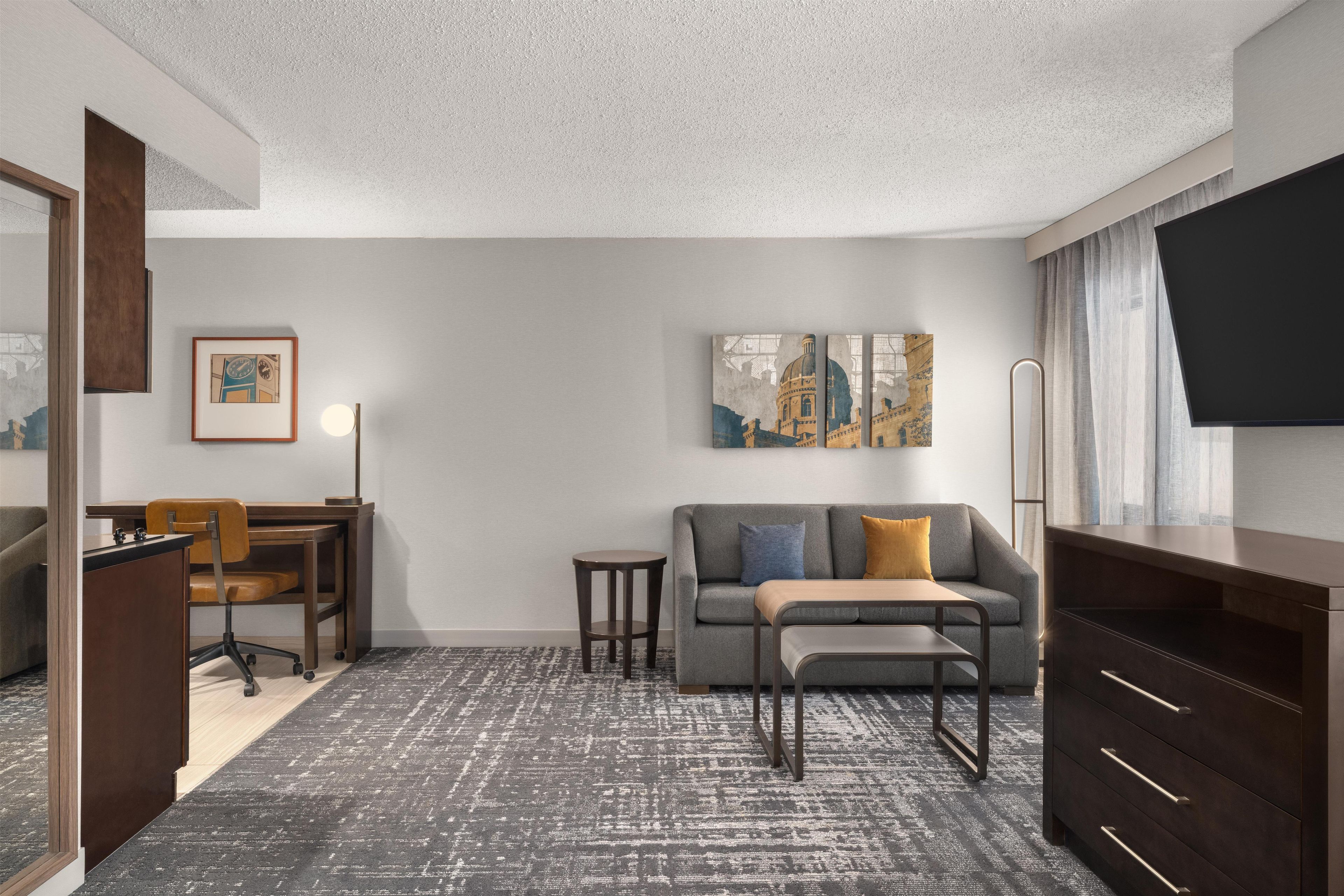 A hotel room with a desk, chair, sofa, and a TV.