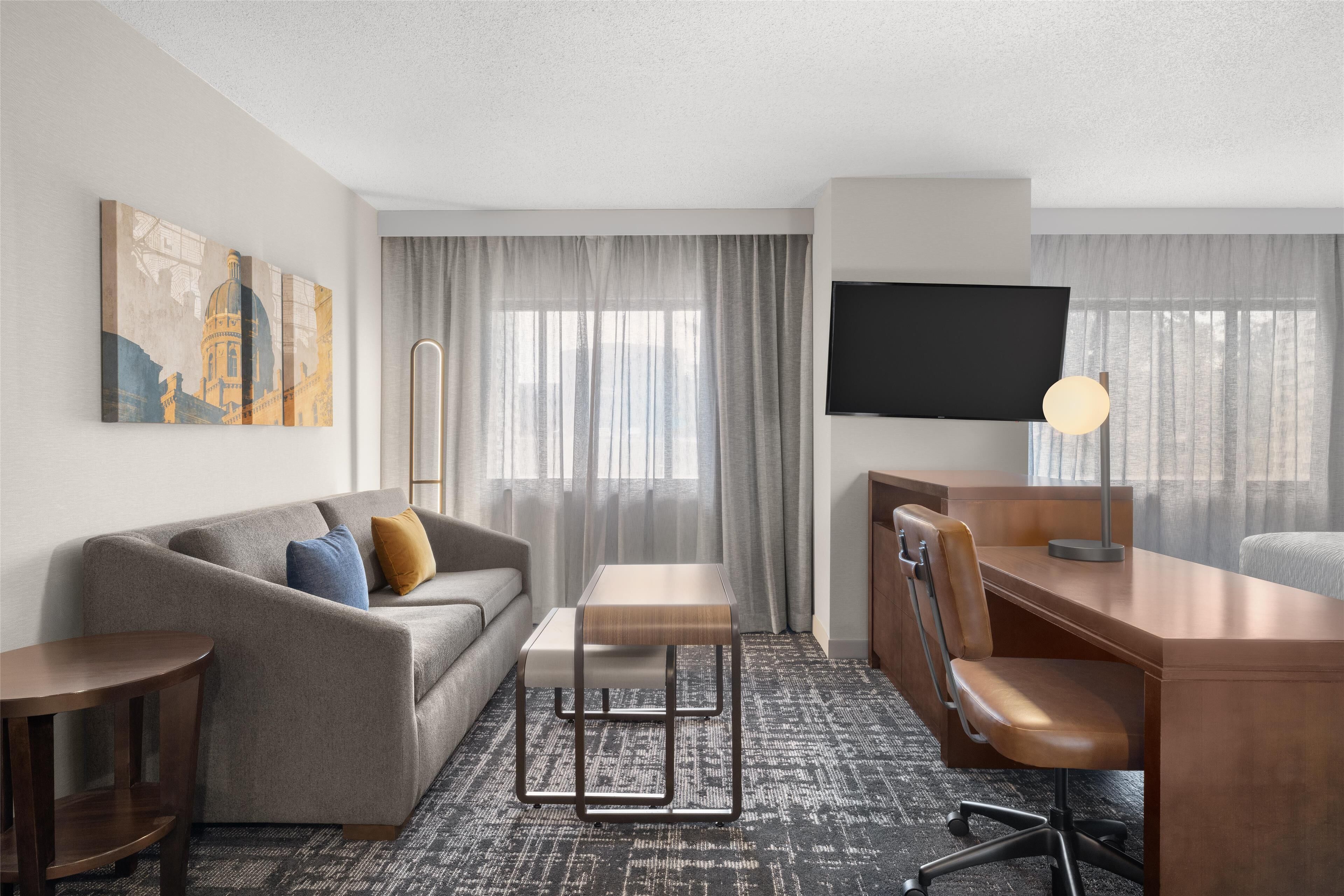 A hotel room with a sofa, a small table, a desk with a chair, a TV, and a large window with curtains.