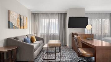 A hotel room with a sofa, a small table, a desk with a chair, a TV, and a large window with curtains.