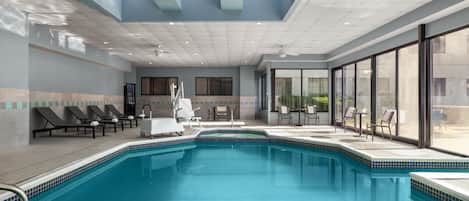 A swimming pool with lounge chairs and a sunbed, surrounded by large windows and a ceiling with recessed lighting.