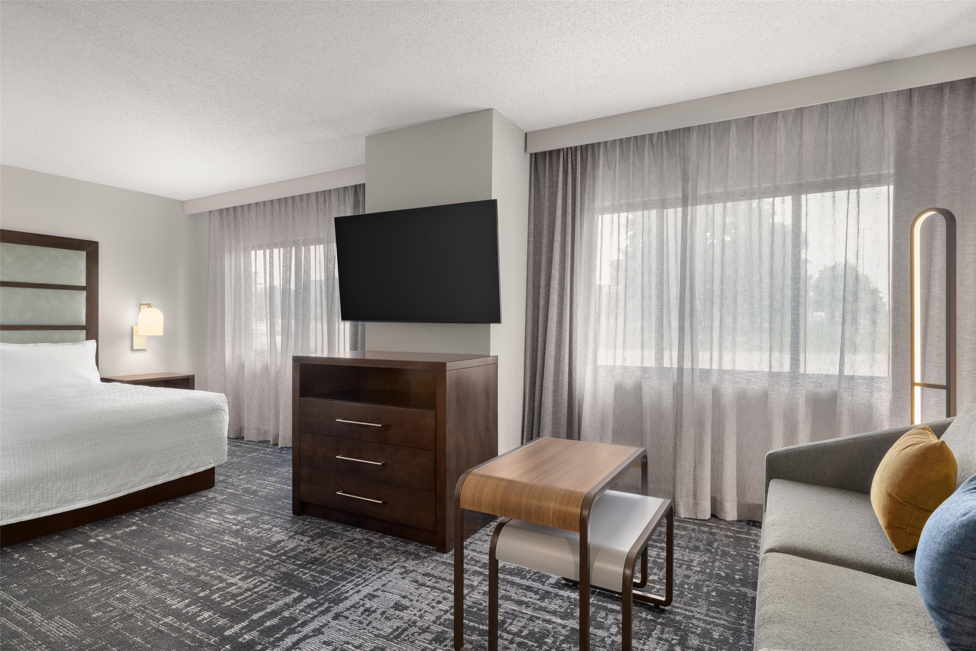 A hotel room with a bed, a TV, a wooden desk, a sofa, and a window with curtains.