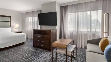 A hotel room with a bed, a TV, a wooden desk, a sofa, and a window with curtains.