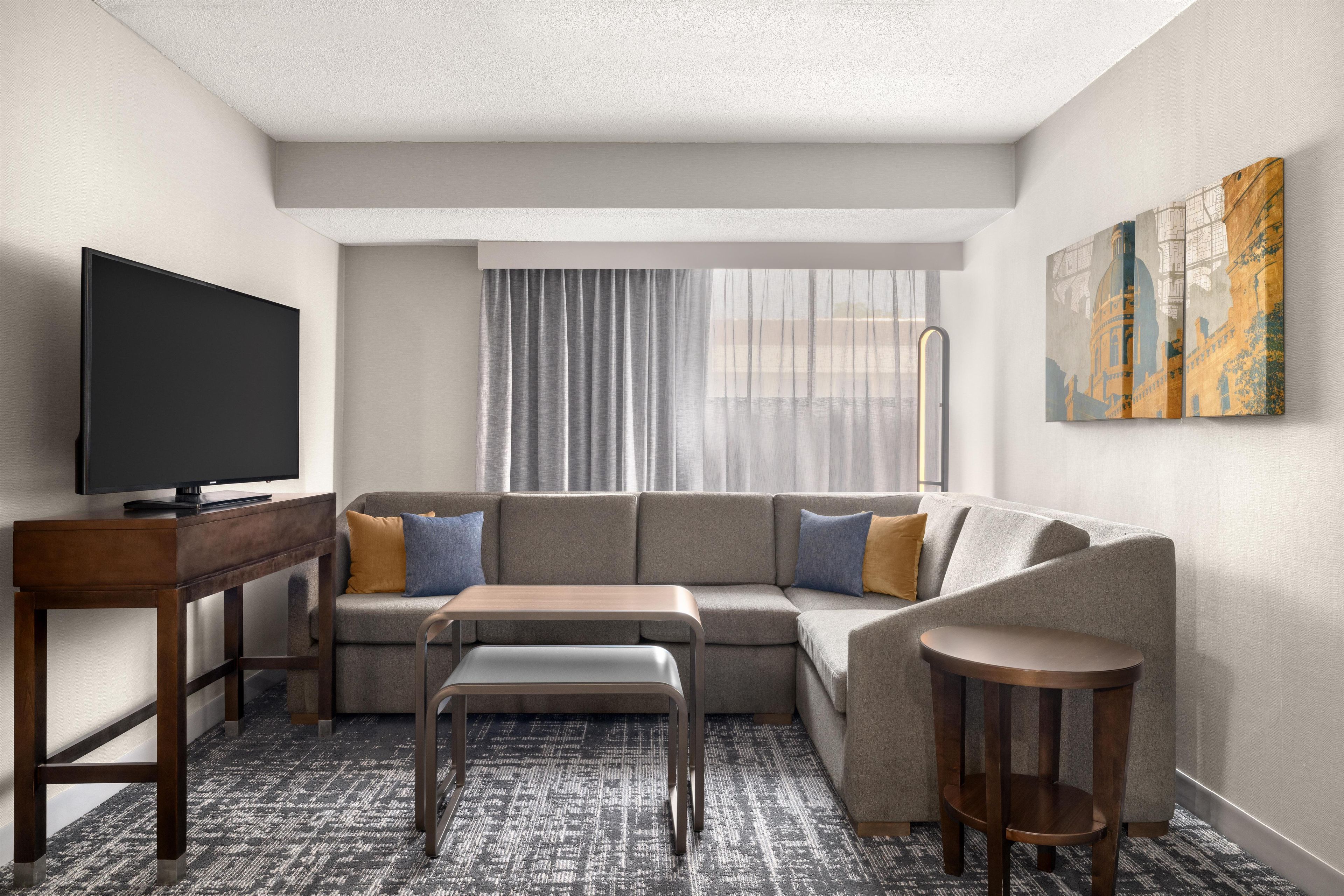 A modern hotel room with a sectional sofa, a flat-screen TV, a small table, and a single chair.