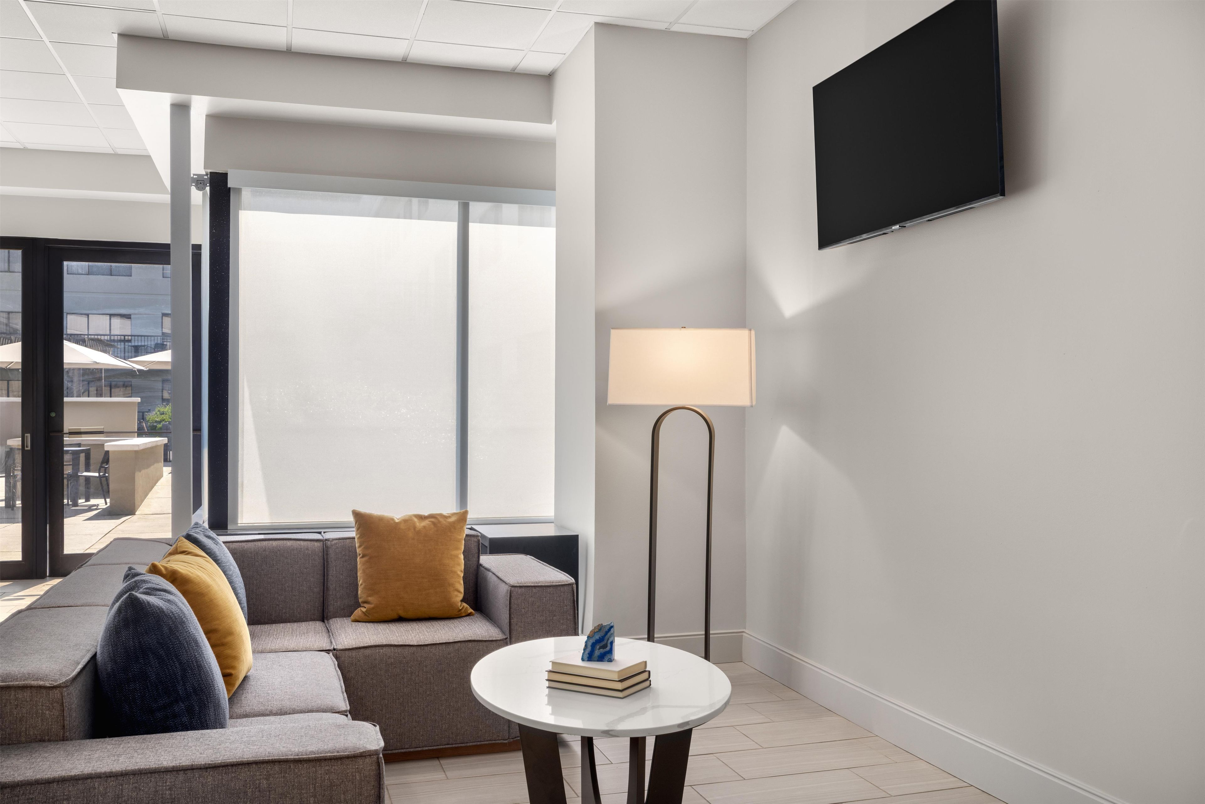 A modern hotel room with a sofa, a small round table with books, a floor lamp, and a TV mounted on the wall.
