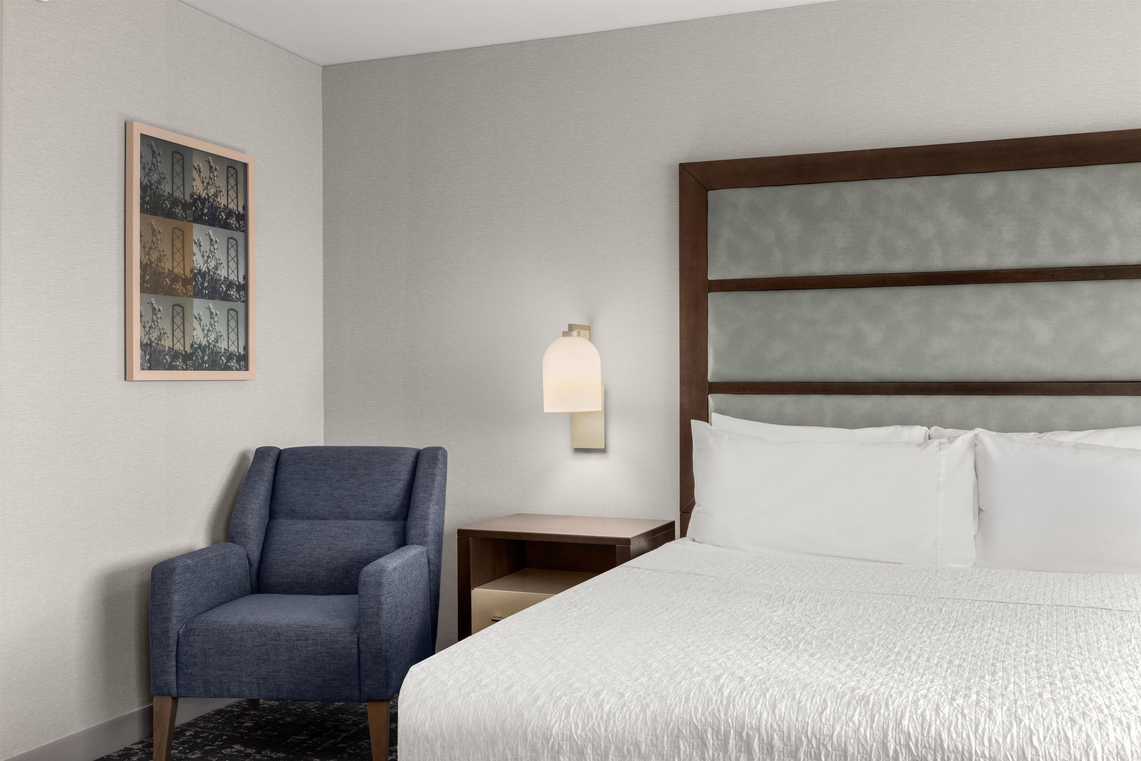 A hotel room with a bed, a chair, a nightstand, a lamp, and a framed picture on the wall.