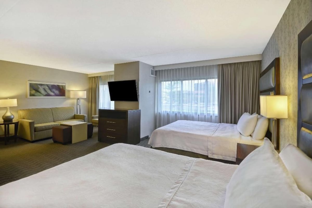 Een hotelkamer met twee bedden, een flatscreen-televisie, een sofa, een bureau en een raam met gordijnen.