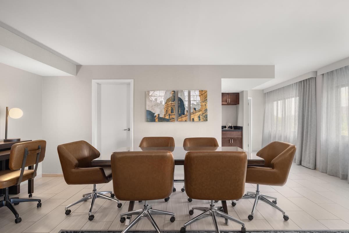 Een vergaderruimte met een witte tafel en zes bruine stoelen, een schilderij aan de muur en uitzicht op een gebouw door het raam.