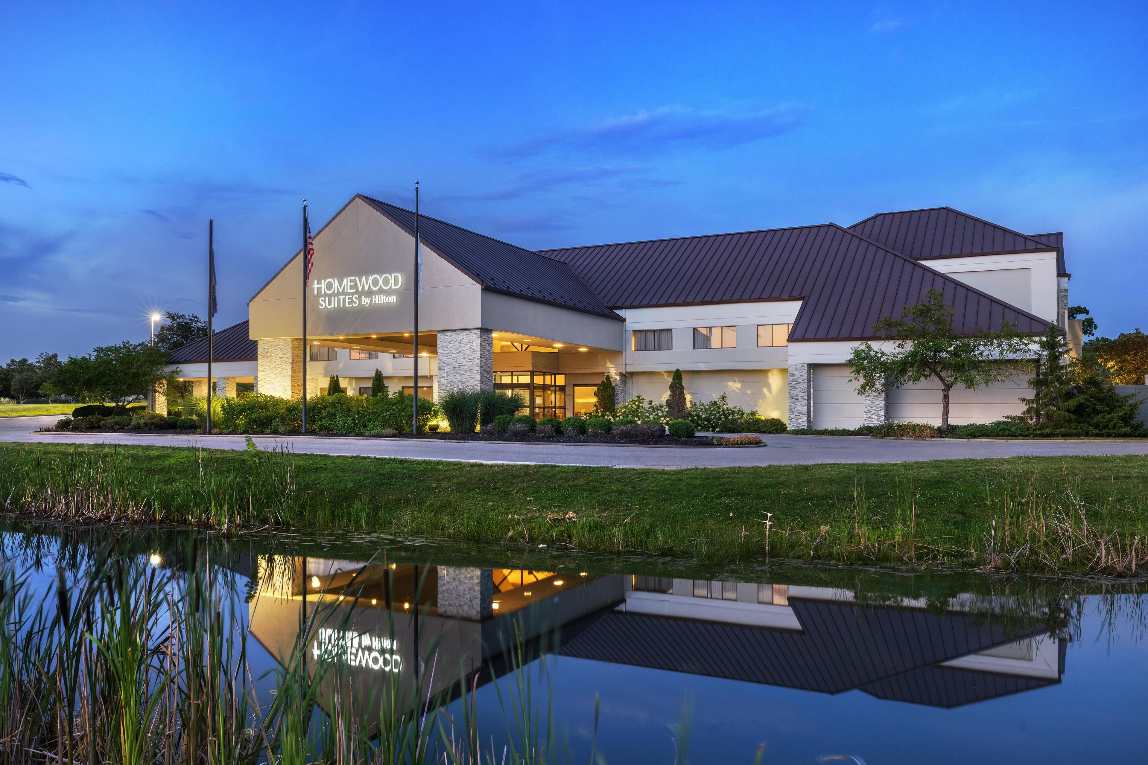 Egy modern stílusú szálloda, melynek homlokzatán nagy méretű „HOMEWOOD SUITES by Hilton” felirat található, és amely egy előtte elterülő tóban tükröződik.