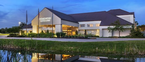 A hotel with a modern design, featuring a large sign for "HOMEWOOD SUITES by Hilton" and a reflection in a pond in front.