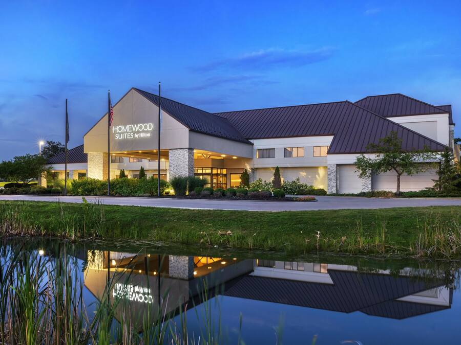 Ein Hotel mit modernem Design, mit einer großen Schilder für "HOMEWOOD SUITES by Hilton" und einem Spiegelbild in einem Teich davor.