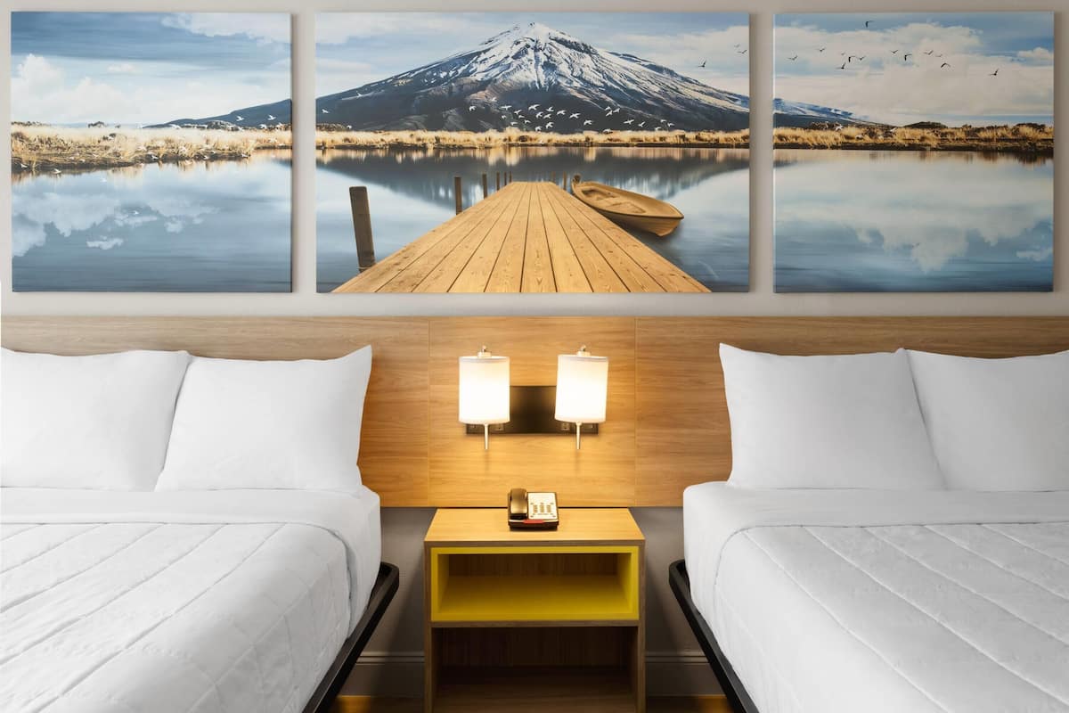 A hotel room with two beds, a wooden nightstand with a phone, and a wall-mounted light fixture.