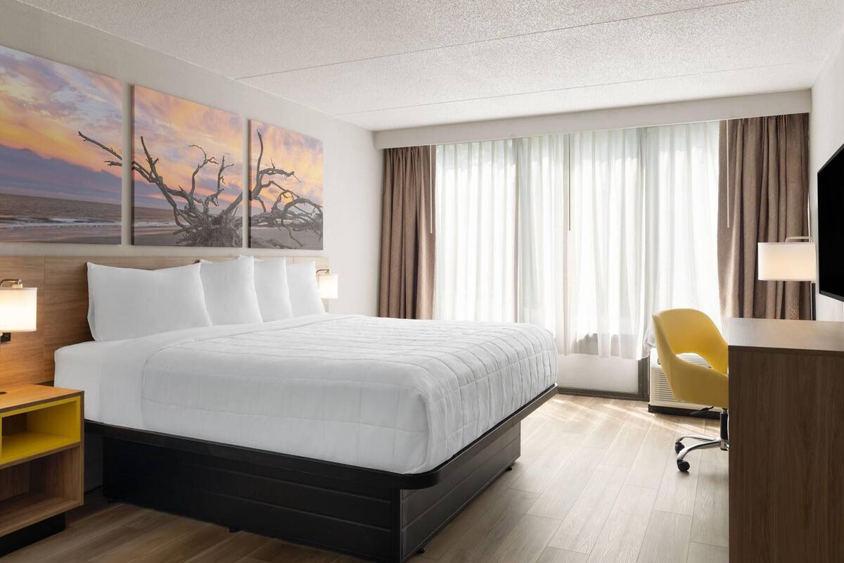 A hotel room with a large bed, a desk with a chair, a TV, and a painting of a sunset on the wall.