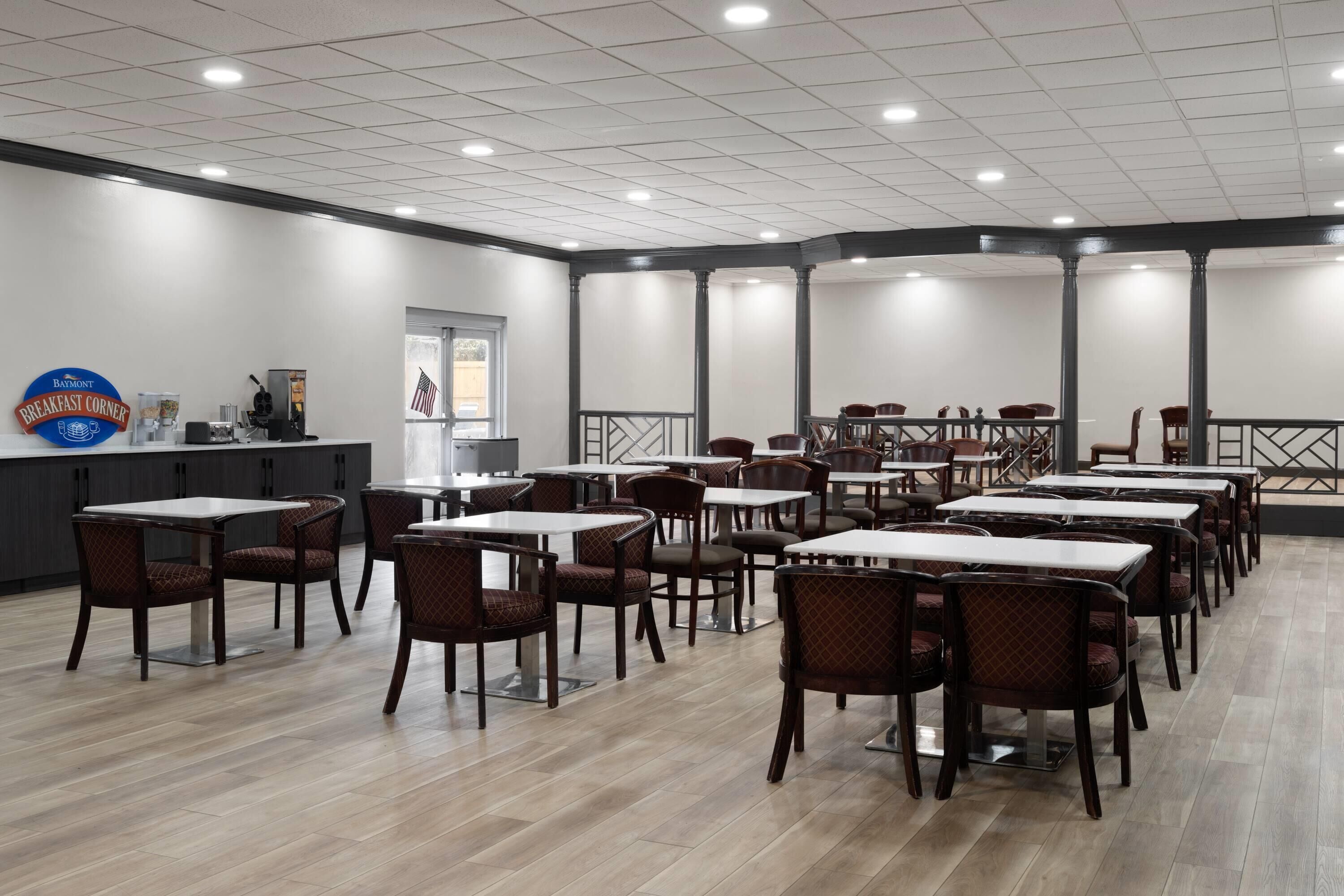 Uma ampla sala de jantar com várias mesas e cadeiras, um balcão de recepção e um sinal indicando um espaço para o café da manhã.