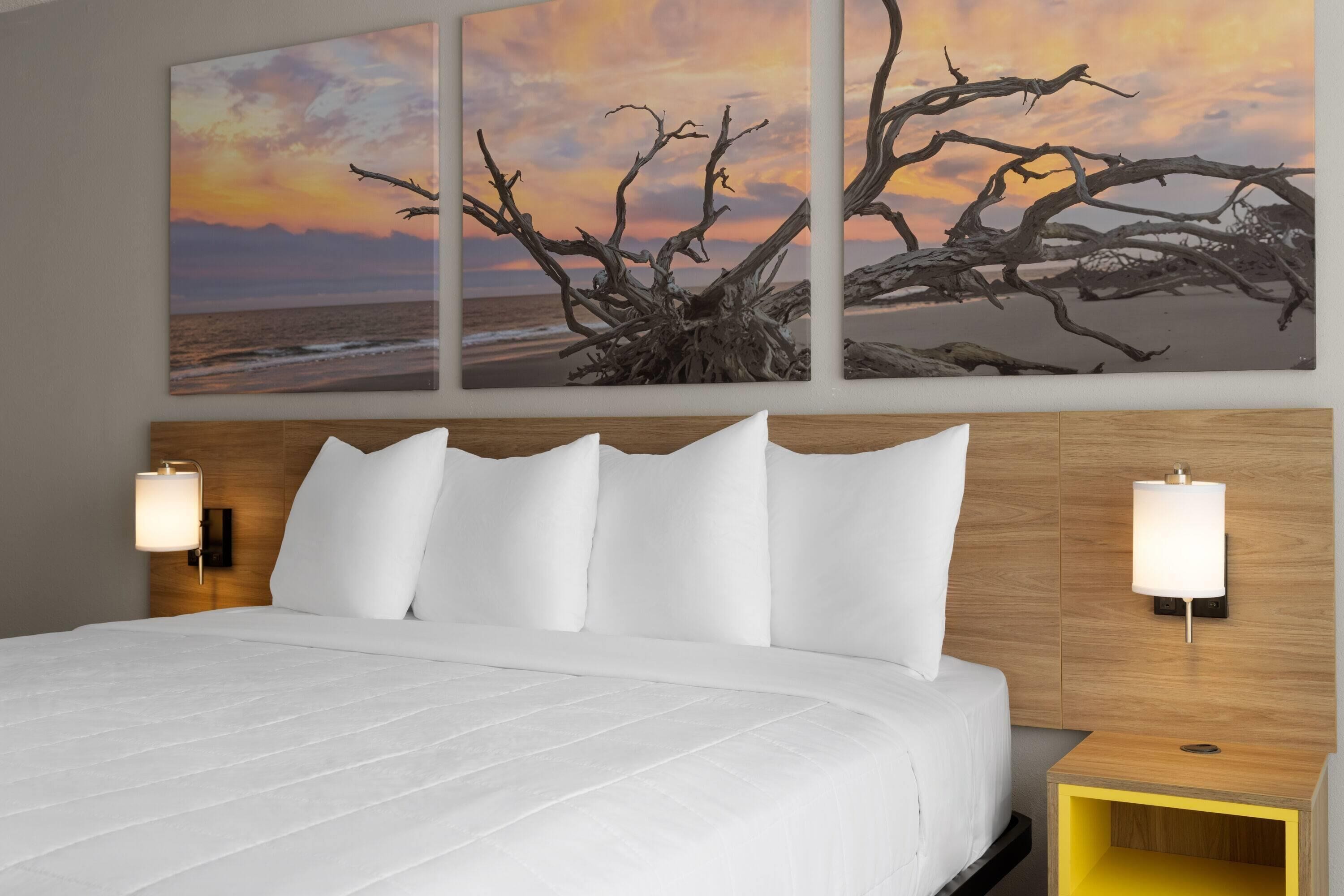 A hotel room with a large bed, two bedside lamps, a wooden headboard, and a wall-mounted artwork of a tree against a sunset.