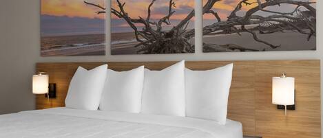 A hotel room with a large bed, two bedside lamps, a wooden headboard, and a wall-mounted artwork of a tree against a sunset.