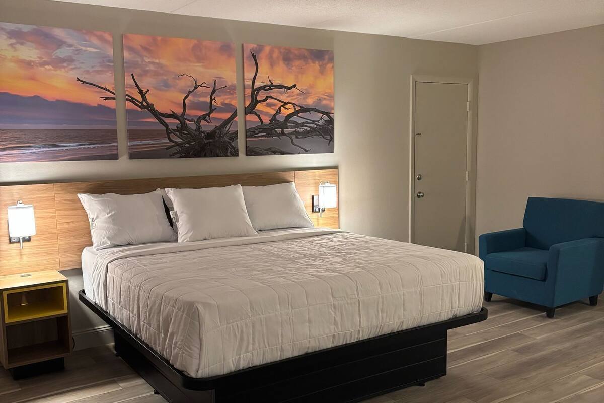 A modern bedroom with a large bed, a nightstand, a blue armchair, and sunset-themed wall art.