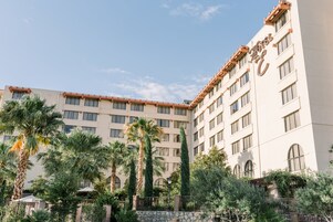 Un hotel de varios pisos con fachada color beige, múltiples ventanas y una pared de piedra con vegetación al frente.