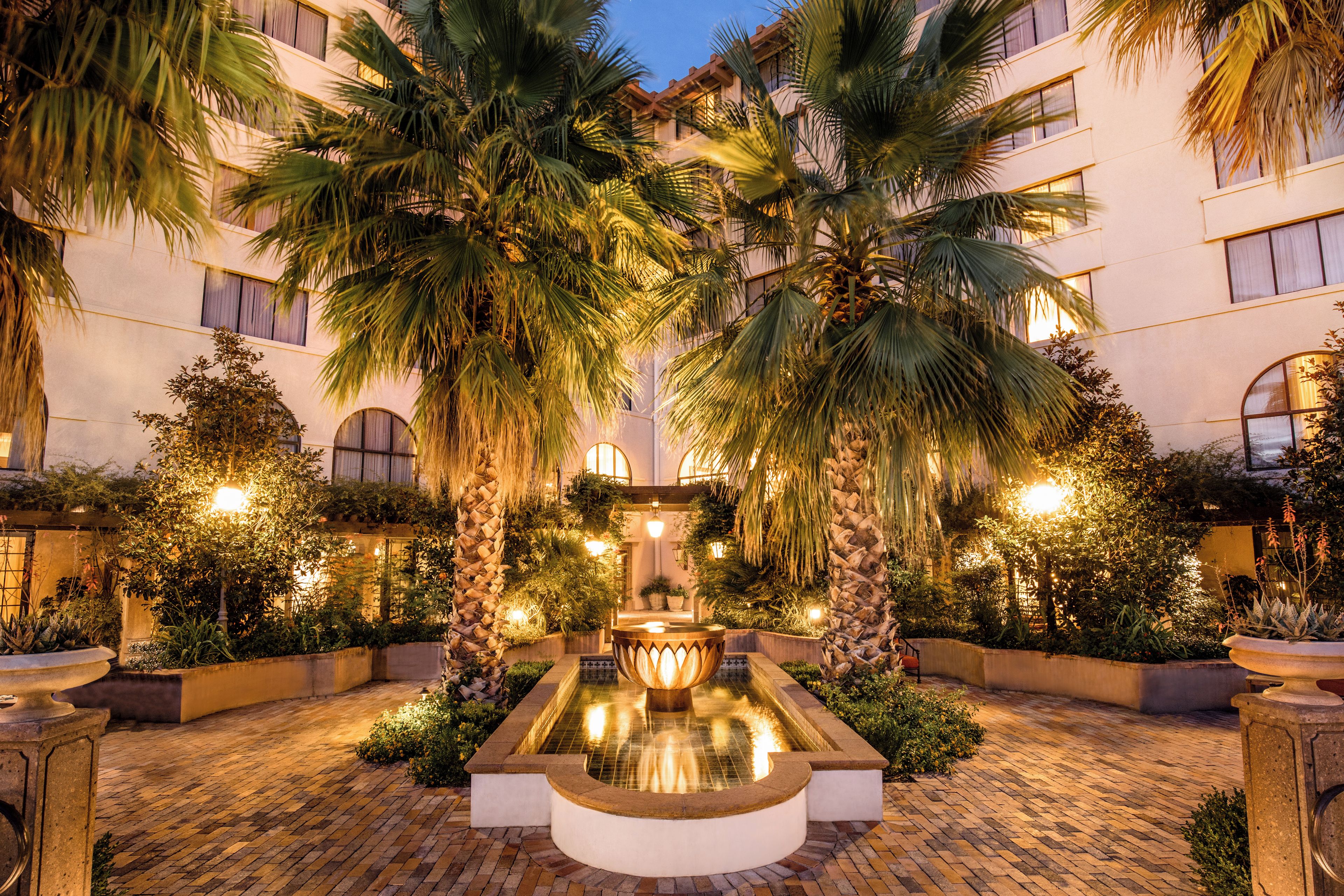Un cortile con una fontana al centro, circondato da palme e illuminato da luci soffuse.