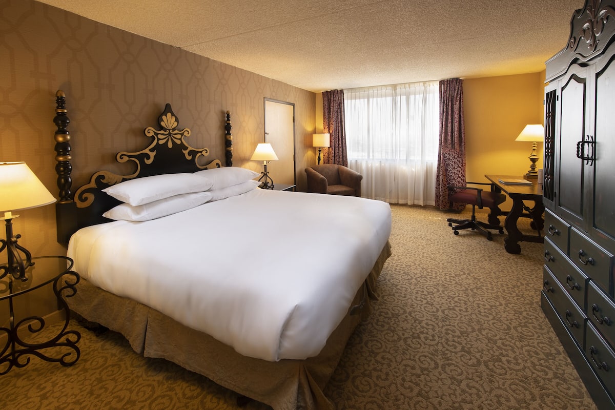 A hotel room with a large bed, two bedside tables with lamps, a desk with a chair, a wardrobe, and a window with curtains.